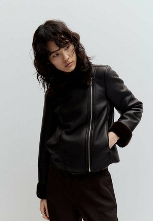 Mujer con cabello oscuro hasta los hombros lleva una chaqueta de cuero negra con cuello y puños de piel, de pie con una mano en el bolsillo.