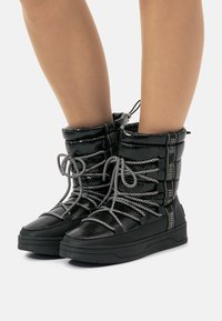 Botas de tobillo acolchadas negras con acabado brillante, que presentan suelas gruesas negras, cordones grises gruesos y parte superior con cordón ajustable para un ajuste personalizable.