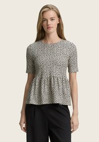 Schwarz-weiß gepunktetes Peplum-Top aus leichtem Stoff, mit kurzen Ärmeln und rundem Ausschnitt. Kombiniert mit schwarzen Hosen.