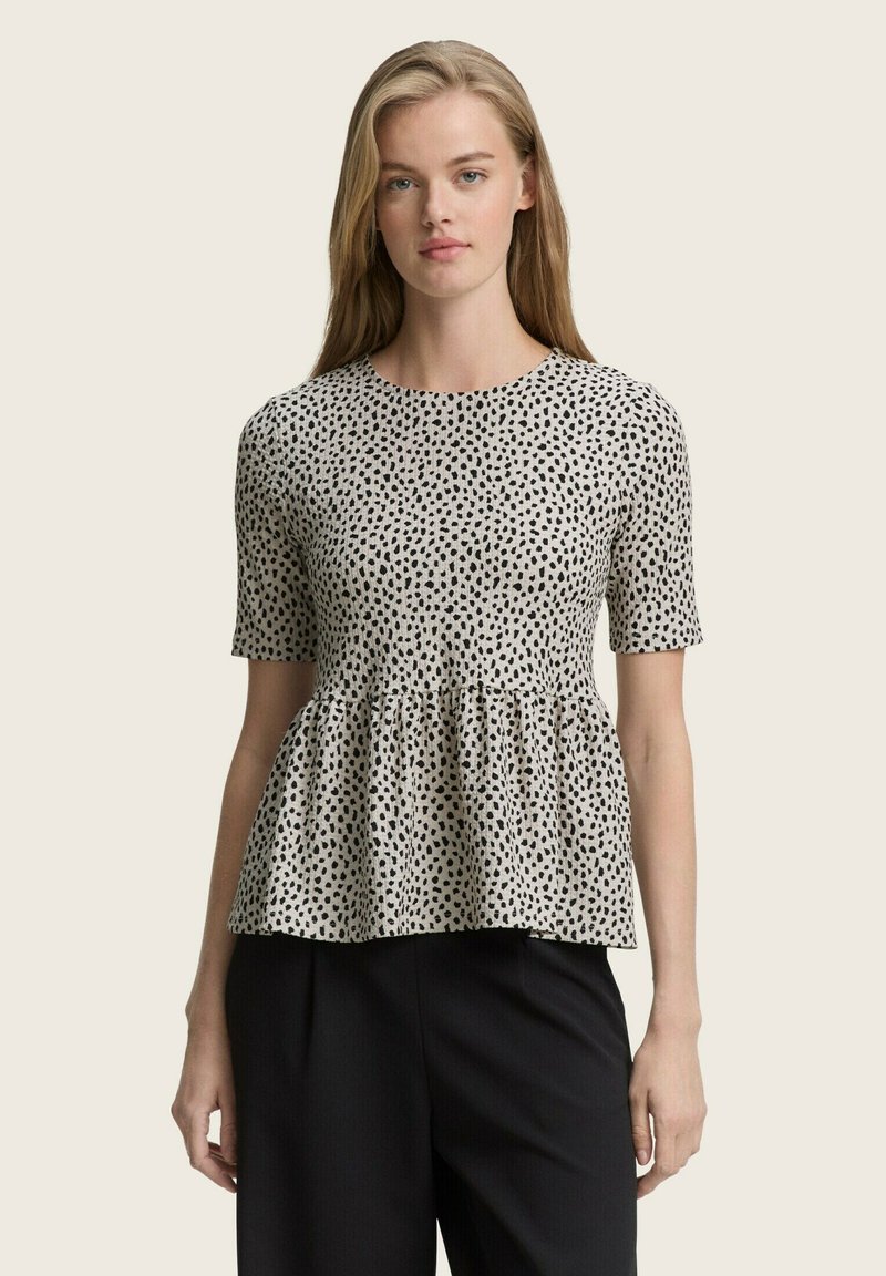 Schwarz-weiß gepunktetes Peplum-Top aus leichtem Stoff, mit kurzen Ärmeln und rundem Ausschnitt. Kombiniert mit schwarzen Hosen.