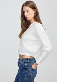 Młoda kobieta z długimi brązowymi włosami nosi białą, krótką bluzę i niebieskie dżinsy, pozując bokiem z jedną ręką w kieszeni na tle jednolitego tła.