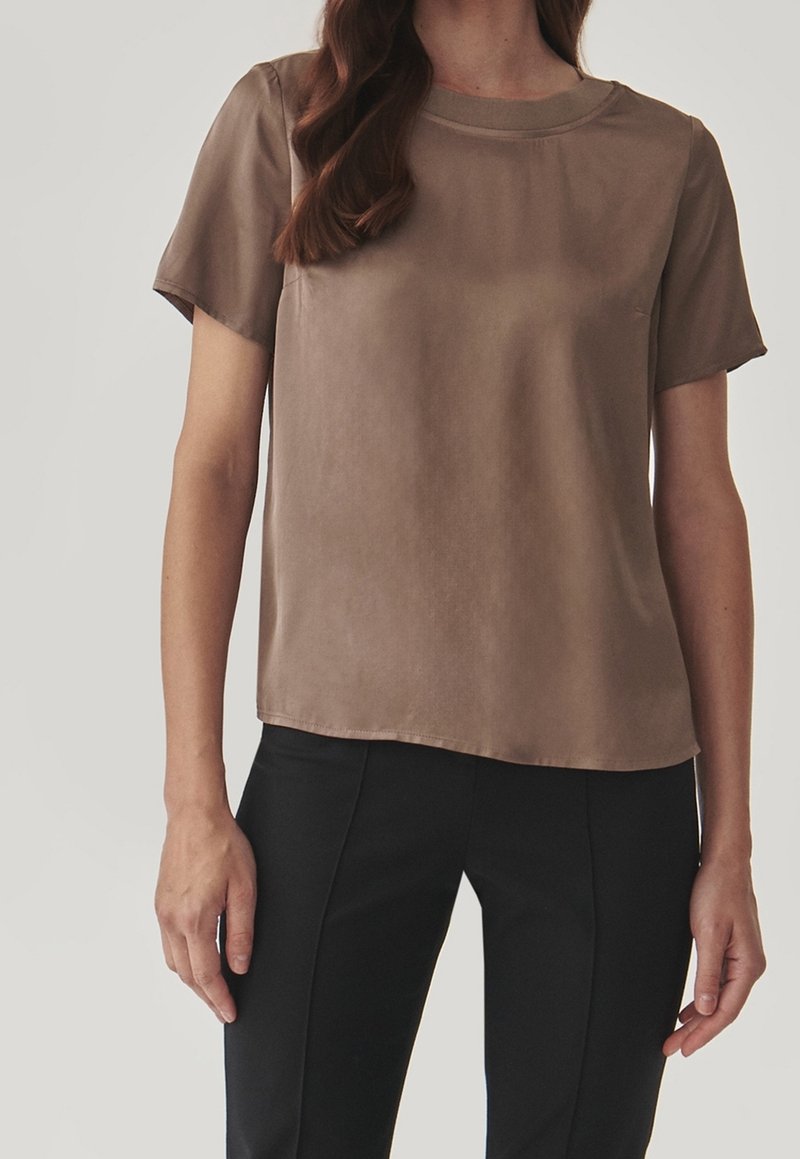 Blouse en soie marron à manches courtes avec un col rond et une texture lisse et légèrement brillante. Associée à un pantalon noir sur mesure.