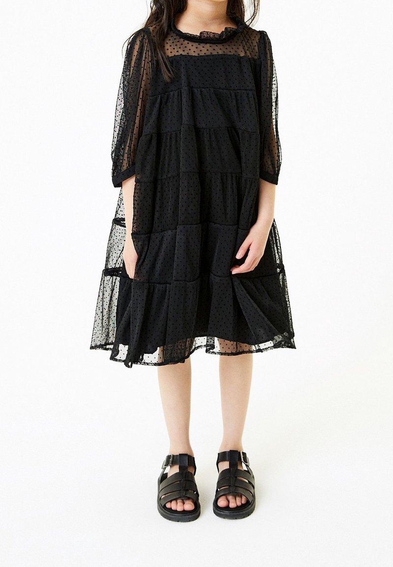 Robe noire à pois avec un design en couches, tissu transparent, col à volants et manches trois-quarts. Associée à des sandales noires à lanières.