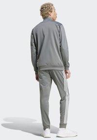 Šedý teplákový súprava so zapínacou bundou a zodpovedajúcimi nohavicami. Obsahuje biele pruhy po stranách a hladký, ľahký materiál.