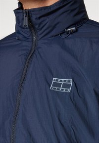 Marinblå jacka med hög krage, med en frontdragkedja och en liten broderad "Tommy Jeans"-logotyp på bröstet. Lätt och strukturerad material.