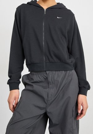 Personne portant un sweat à capuche zippé noir avec un petit logo blanc et un pantalon cargo ample gris foncé avec des poches latérales.