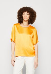 Top corto in satin a maniche corte di un vivace arancione con scollo rotondo. Presenta una vestibilità ampia e una texture liscia, abbinato a pantaloni chiari.