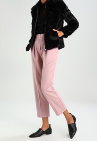 Giacca in pelliccia sintetica nera con zip frontale, abbinata a pantaloni rosa chiaro su misura e corti, e ballerine nere con punta. Texture lisce, vestibilità moderna.