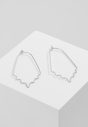 Boucles d'oreilles en fil métallique ton argent avec des bords inférieurs en zigzag angulaire, présentées sur un piédestal géométrique blanc.