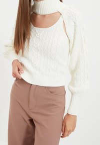 Biały sweter o splocie warkoczowym z wysokim kołnierzem i ściągaczem u dołu. Posiada bufiaste rękawy oraz małe ozdobne guziki z przodu. Wyeksponowane brązowe spodnie.