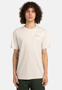 Camiseta de algodón beige con cuello redondo y mangas cortas. Presenta un sutil texto verde: "Element Skateboard Co." en el lado izquierdo del pecho.