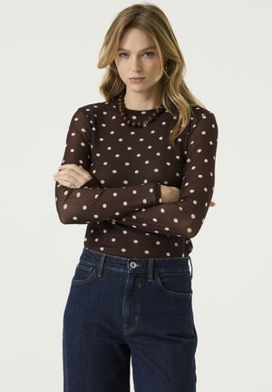 Frau mit blondem Haar, trägt ein dunkelbraunes langärmliges Polka-Dot-Oberteil, blaue Jeans und eine dicke braune Perlenkette, Arme verschränkt.