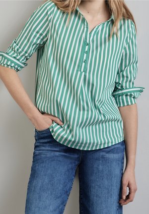 Kobieta w zielono-białej koszuli w pionowe paski, z podwiniętymi rękawami, i niebieskich dżinsach, z ręką w kieszeni.