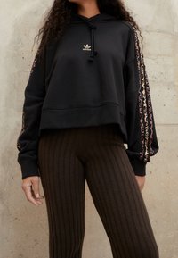 Felpa corta nera con cappuccio e lacci, con logo dorato, caratterizzata da strisce a motivo animale lungo le maniche. Abbinata a leggings marroni a coste.