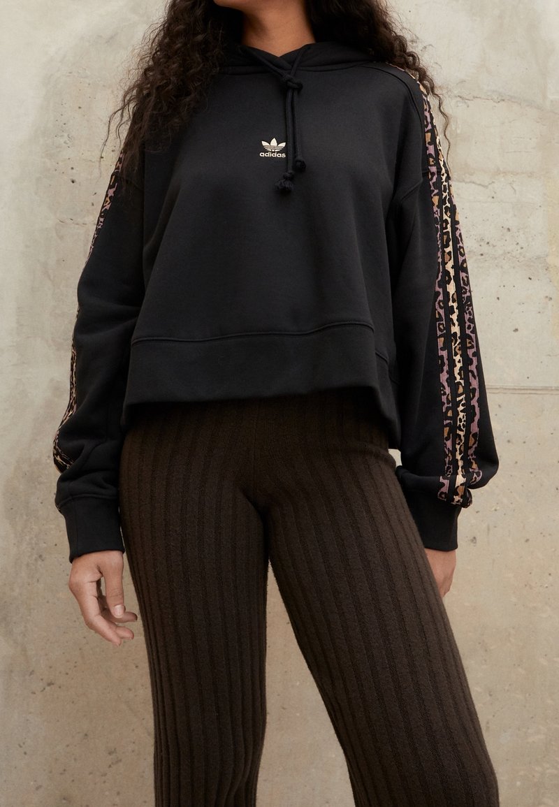 Felpa corta nera con cappuccio e lacci, con logo dorato, caratterizzata da strisce a motivo animale lungo le maniche. Abbinata a leggings marroni a coste.