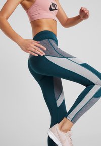 Tyrkysové sportovní legíny s šedými a světle šedými panely; mají vysoký pas a elastickou tkaninu; doplněné světle růžovými sportovními botami.