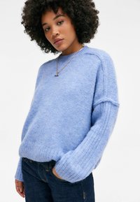 Helles blauer, gestrickter Pullover mit einer weichen, flauschigen Textur, gerippten Bündchen und einem lockeren Schnitt. Kombiniert mit dunklen Jeans.