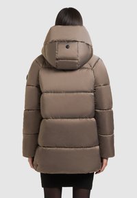 Woman wears taupe puffer jacket with large hood, viewed from the back, standing against plain light background.