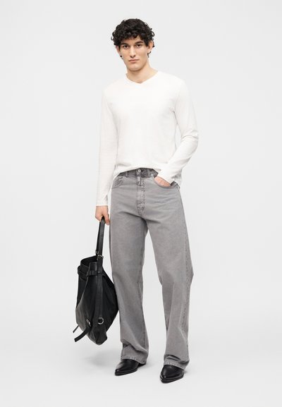 Νέος άνδρας με σγουρά μαλλιά, φορώντας λευκό πουκάμισο μακρυμάνικο, γκρι τζιν φαρδιάς γραμμής, μαύρα μποτάκια, κρατώντας μαύρη δερμάτινη τσάντα σε λευκό φόντο.