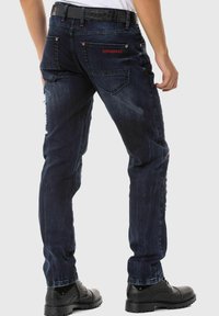 Dunkelblaue Denim-Jeans mit einem abgenutzten Finish, sichtbaren Nähten und roten Akzenten. Ein schwarzer Gürtel ergänzt das Design. Slim-Fit-Stil.