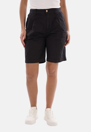Personne portant un short noir au genou avec un bouton et des plis, des baskets blanches et un haut blanc, debout droite sur un fond uni.