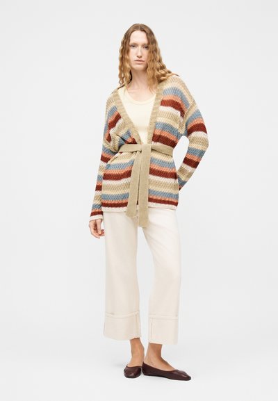 Γυναίκα που στέκεται φορώντας μπεζ παντελόνι, ριγέ πλεκτή ζακέτα με ζώνη, κρεμ τοπ και καφέ επίπεδα παπούτσια σε απλό φόντο.