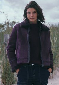 Giacca viola in finta suede con colletto e polsini in pile, chiusura con zip e tasche laterali, indossata sopra un maglione bordeaux a coste e jeans scuri.