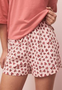 Lyserøde bomuldsshorts med elastisk talje, prydet med et spredt hjerte-mønster i forskellige nuancer af rød. Blødt, afslappet design.
