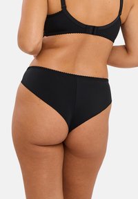 Femme portant un soutien-gorge noir et des culottes taille haute, montrant son dos et un côté partiel contre un fond uni.