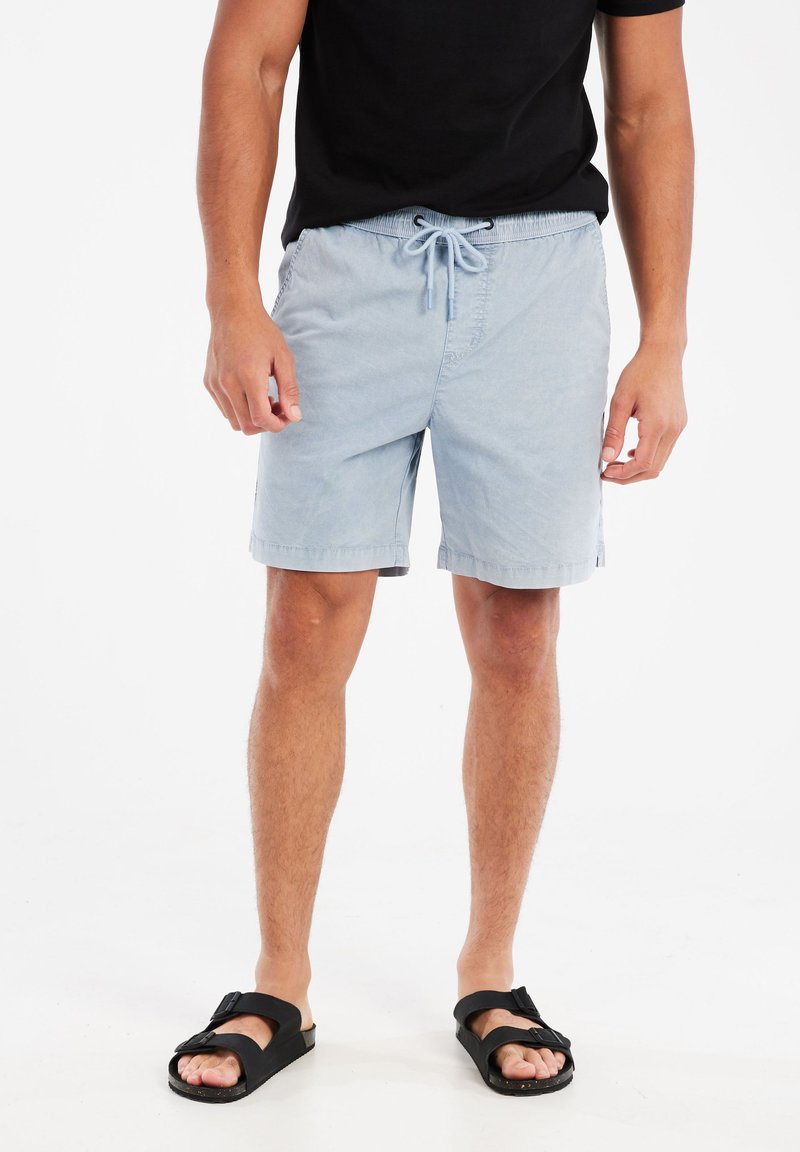 Lichtblauwe zwembroek met een elastische tailleband en een verstelbaar trekkoord. Voorzien van zijzakken en een relaxte pasvorm. Gekoesterd met zwarte sandalen.