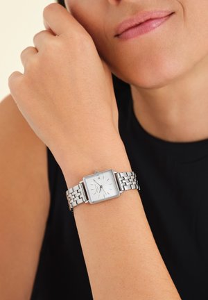 Montre en acier inoxydable argenté avec un cadran blanc carré, marqueurs d'heure minimalistes et un bracelet à maillons, portée sur un poignet contre un haut noir.
