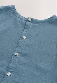 Tissu en lin bleu clair avec un col rond simple et une rangée verticale de quatre boutons blancs au centre.