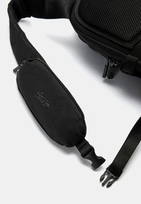 Sort skuldertaske med en tekstureret rem, der har en lille lynlåslomme. Holdbart stof med en glat overflade og matte hardware-detajler.