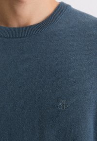 Niebieski sweter z prążkowanym, okrągłym dekoltem, wykonany z miękkiej tkaniny. Na piersi znajduje się małe, haftowane logo w dopasowanym odcieniu niebieskiego.