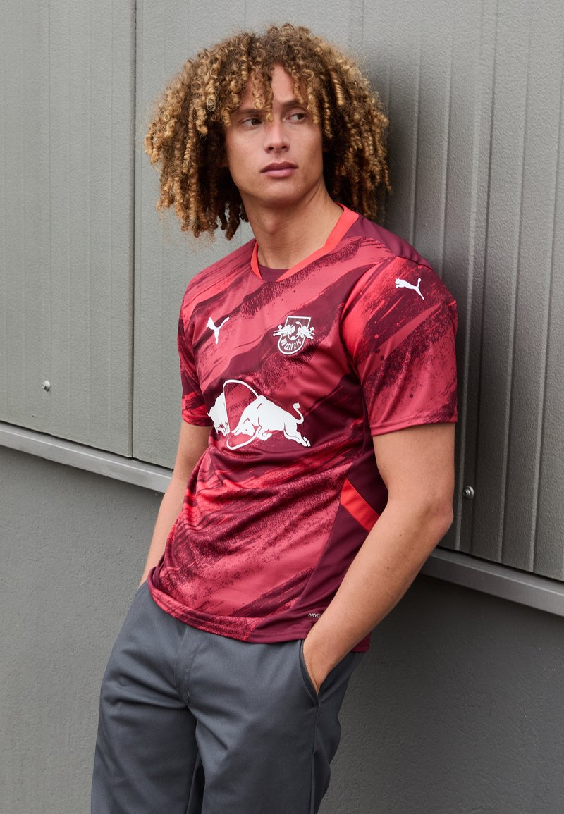 Maglia sportiva rossa e nera con motivi astratti, con un logo bianco sul petto. Il design include maniche corte e scollatura rotonda.
