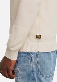 Maglione lavorato a maglia beige con polsini e orlo a coste, caratterizzato da un'etichetta nera e gialla sul lato. Indossato con jeans azzurri chiari.