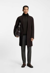 Długi brązowy płaszcz na ciemnym golfie, szare spodnie w wzór i brązowe loafersy. Trzyma ciemną skórzaną torbę z paskiem na ramię.