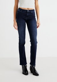 Temno pranje jeans hlače s ravnim krojem, z zapiranjem na gumb in dvema sprednjima žepoma. Združene z črnimi gležnjarji.