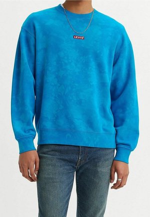 Persona usando un suéter Levi's teñido con efecto tie-dye azul, collar de cadena plateada, anillo de plata y jeans azules, de pie frente a un fondo blanco liso.