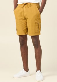 Pantalones cortos cargo caqui con múltiples bolsillos laterales, cintura con cordón y una textura en la tela. Llevados con zapatillas blancas sobre un fondo neutro.