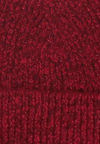 Beanie en tricot rouge avec un motif texturé, présentant un revers replié et un ajustement snug. Le matériau semble doux et extensible.