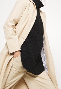 Beige trench coat over a black knitted turtleneck and light beige pants. The coat has a relaxed fit, and the turtleneck features a textured pattern.