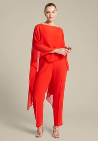 Rode twee-delige outfit met een vloeiende, doorschijnende top met vleermuismouwen en aangesloten broeken, aangevuld met strakke metallic hoge hakken.