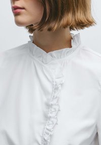 Woman with short light brown hair wearing a white blouse with a ruffled stand collar and ruffle trim along the front.