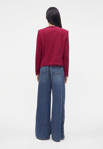 Femme debout, dos à l'appareil photo, portant une veste rouge texturée à ourlet effiloché et un jean bleu à jambes larges sur fond uni.