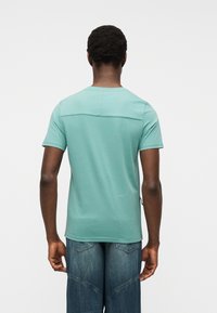 Άνδρας με ανοιχτό τιρκουάζ κοντομάνικο t-shirt και μπλε τζιν παντελόνι, εμφανιζόμενος από πίσω με φόντο απλό λευκό.