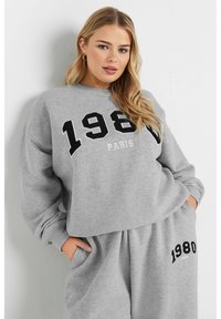 Sudadera gris con bordado negro de "1980" y "PARÍS", puños acanalados y un corte relajado. Combinada con pantalones de chándal grises a juego.