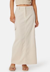 Beige maxi-jurk met een rechte snit, zijzakken en een gladde, textuurstof. Draagt met sierlijke platte sandalen.