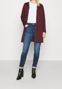 Kvinna i mörkröd lång cardigan, vit skjorta, uppvikta blå jeans och svarta klackade ankelstövlar som står mot vit bakgrund.