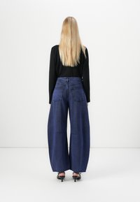 Wide-leg dark blue denim pants with a high waist, textured fabric, and two back pockets, paired with a black long-sleeve top and heels.